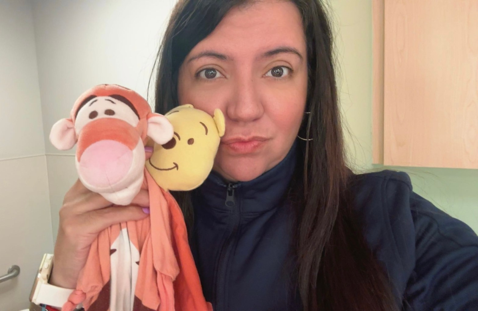 Persona de cabello largo y oscuro, con chaqueta azul marino, sostiene junto a su rostro un peluche de Winnie the Pooh y una mantita de apego de Tigger.